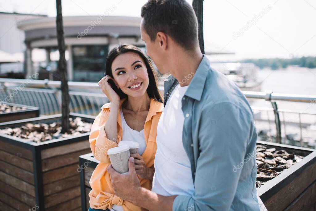 Smiling young Asian woman looking with love and tenderness at boyfriend while standing together on river bank in city talking and drinking coffee