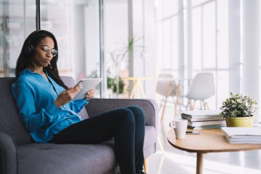 Gözlüklü, kendinden emin ve başarılı Afro-Amerikan çalışanın parlak ofisinde tabletle oturup kulaklıkla müzik dinlemesinin yan görüntüsü.