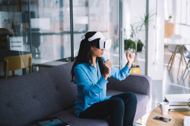 VR kulaklık takmış, kanepeye oturmuş sanal bir objeyi elinde tutuyor gibi el kol hareketi yapan genç bir kadının yan görüntüsü.