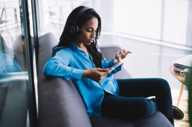 Mavi gömlekli, koyu renk pantolonlu, gri koltukta oturan ve şeffaf elektronik aletlere dokunan siyahi bayanın yan görüntüsü.