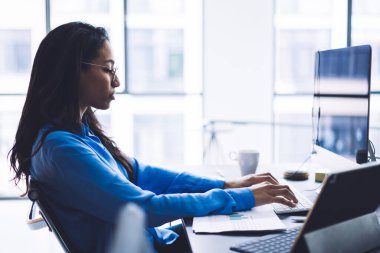Afrikalı Amerikalı kadın ofis memurunun bilgisayar kullanarak rapor verirken ve çalışma alanındaki grafiklerin çıktısını alırken yan görünüm