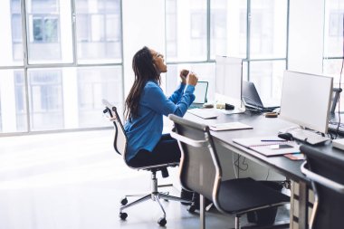 Açık modern uzay ofisinde bilgisayar başında otururken ve el kaldırırken çığlık atan resmi giyinmiş ve gözlüklü mutlu Afrikalı Amerikalı kadın.