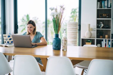 Gülümseyen Kafkas kadın dijital göçebe dairede oturuyor serbest çalışmaktan memnun, mutlu 20 'li yaşlarda yeniyetme kız çevrimiçi kurs öğreniyor ve dizüstü bilgisayarında webinar eğitimi izliyor.