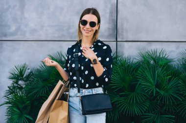 Moda kıyafetli ve güneş gözlüklü neşeli genç bir kadın. Yeşil torbaların önünde durmuş gülümsüyor ve kameraya bakıyor.