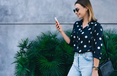 Gayri resmi giysiler içinde, zengin bitkilerin önünde dikilirken sosyal medyada gezinen neşeli genç bir kadın.