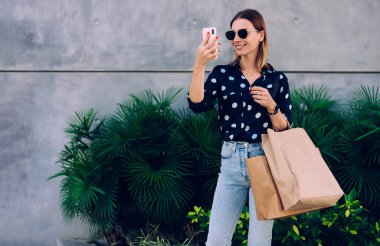 Güneş gözlüklü, şık gülümseyen genç bayan alışveriş merkezinin yanında elinde kese kağıtlarıyla duruyor ve cep telefonuyla sosyal medya için selfie çekiyor.