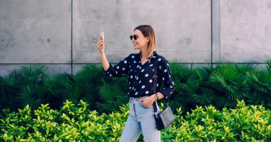 Güneşli bir günde yeşil tropikal bitkiler tarafından çerçevelenmiş taş duvarın önünde dururken, güneş gözlüklü ve günlük giysili güzel bir bayanın akıllı telefonuyla selfie çekişi.