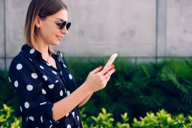 Güneşli bir günde yeşil bitkilerin yanında yürürken ve ekrana bakarken günlük kıyafetli, güneş gözlüklü, akıllı telefonlu genç bir kadının yan görüntüsü.