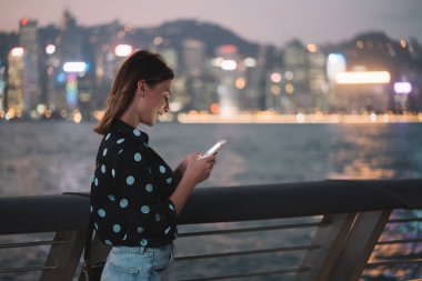 Akşamları şehir manzarası olan sette yürürken akıllı telefon kullanan sıradan kıyafetli mutlu genç bir kadının yan görüntüsü