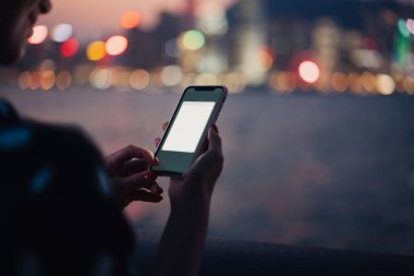 Plastik çantada modern akıllı telefon kullanan tanınmayan bir kadın. Uygulama, gece nehri ve Bokeh şehir ışıklarının bulanık arkaplanını taklit ediyor.