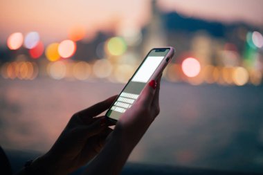 Pembe çantada modern akıllı telefonu kullanan tanınmayan bir kadın sürüsü kuryeyle birlikte gece nehrinin bulanık arka planında ve Bokeh şehrinin ışıklarında makara yapıyor.