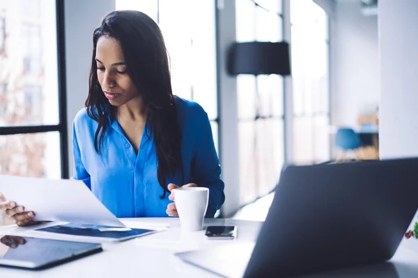 Serious calm African American adult female in formal clothes checking papers while sitting at table with digital devices and white cup with hot drink against big window in light modern loft office