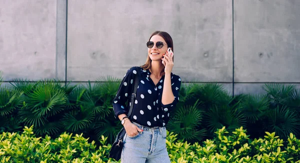 穿着休闲装 戴着太阳镜 手持手提包的年轻女子一边站在石墙和绿色热带植物的后面一边一边一边一边一边用手机与人交谈 一边望着远方 — 图库照片