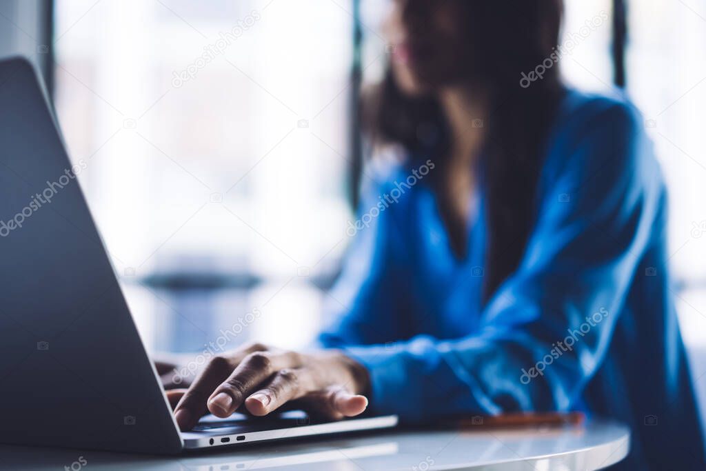 Soft focus of African American female sitting at table in light office and clicking on modern laptop keyboard while working