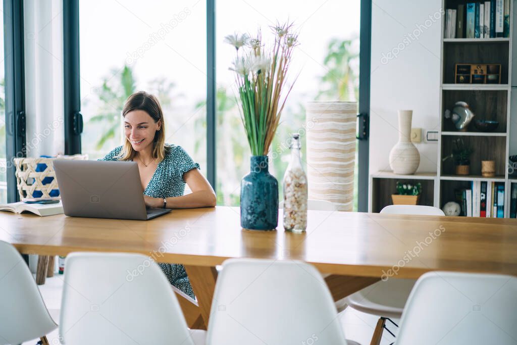Smiling caucasian female digital nomad sitting at table in apartment satisfied with freelance job, happy clever 20s hipster girl learning online course and watching training webinar on laptop computer