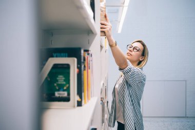 Barselona 'da üniversite kütüphanesinde okurken kitaplıktan kitap alan gözlüklü ve günlük giysili akıllı kadınların yan görüntüsü