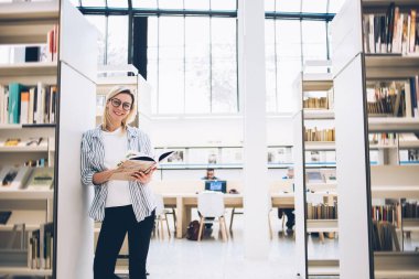 Kitapla gülümseyen ve kameraya bakan neşeli genç bir bayan. Barcelona 'daki modern kütüphanede koridorda durup kitaplığa yaslanırken.