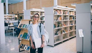 Bilgisayarlı ve sırt çantalı çağdaş akıllı kadın Barcelona 'daki kütüphane çalışmaları sırasında kütüphanelerde dururken kameraya bakıyor.