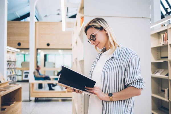 Glad Young Woman Glasses Leaning Bookshelf Reading Interesting Book While — Stock Photo, Image