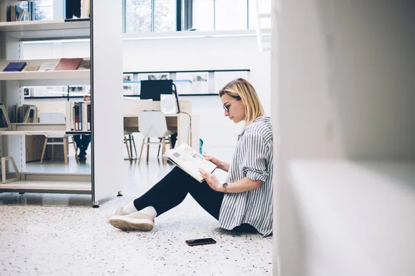 バルセロナ市内の図書館で勉強しながら 本棚の近くの床に座って面白い本を読んでいる眼鏡の賢い女性の側面図 — ストック写真