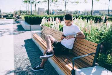 Gündelik modern adam yaz kıyafetleri içinde şehir meydanındaki ahşap bankta takılıyor ve parlak güneş ışığında cep telefonuna bakıyor.