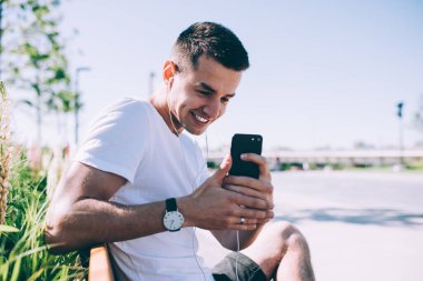 Kulaklık takmış, akıllı telefon kullanarak fotoğraf çeken ve gün ışığında parkta oturmuş müzik dinleyen genç bir adamın yan görüntüsü.
