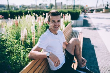 Kulaklıkla konferans dinlerken kameraya gülümseyen yakışıklı erkek öğrenci bir ayağı yukarıda bankta güneşlenmenin keyfini çıkarıyor.