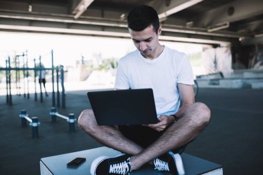 Günlük kıyafetlerle gezen, bacak bacak üstüne atmış ve internette gezen genç bir erkek yaz günü spor salonunda dizüstü bilgisayara odaklanmış.