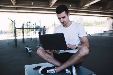 Spor sahasında bacak bacak üstüne atarak otururken, günlük kıyafetleri içinde koyu renk saçlı, laptopta gezinen odaklanmış serbest çalışan bir erkek.