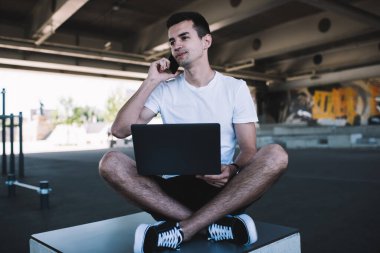 Düşünceli genç erkek bilgisayarını karıştırıyor ve köprü altında bacak bacak bacak üstüne atarak otururken cep telefonuyla konuşuyor ve başka tarafa bakıyor.
