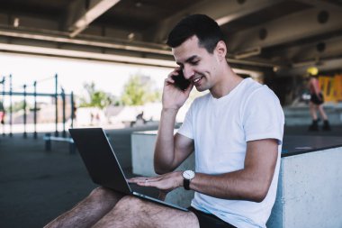 Cep telefonuyla konuşurken ve köprü altında otururken, kol saati olan neşeli genç bir adam netbook 'ta yazıyor.