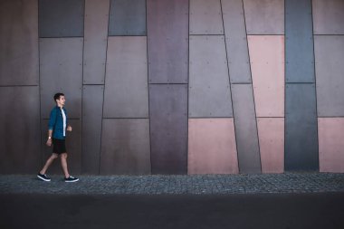 Şehirdeki metalik duvarın yanında yürüyen, günlük kıyafetler ve kol saati takmış spor ayakkabılı ciddi bir gencin yan görüntüsü.