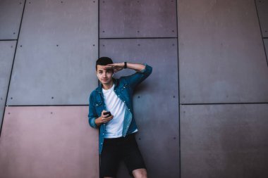 Moda kıyafetleri ve kulaklıkları içinde duvarın yanında duran ve duvara yaslanıp selamlaşırken akıllı telefon kullanan mutlu genç adam.