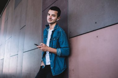 Kot giyen, modern binanın yanında dikilen, akıllı telefon ve kablosuz kulaklık kullanan, İnternet 'te sörf yapan pozitifçi genç bir erkekten.