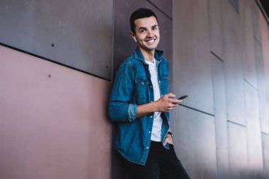 Metal duvarın yanında durup müzik dinlerken ve sokakta kameraya gülümserken gülümseyen modaya uyan genç adam.