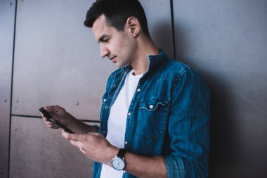 Kot elbiseli, binanın duvarının yanında duran ve uygulamanın kart verileri için akıllı telefon kullanan odaklanmış genç bir erkeğin yan görüntüsü