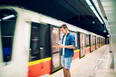 Metro istasyonunda hareket halindeki metro treninin yanında duran kot pantolonlu, cep telefonunu kontrol eden pozitiflerin yan görüntüsü