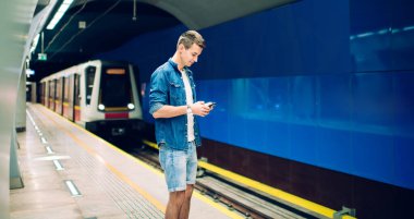 Demiryolu platformunda metro trenini beklerken günlük kıyafetleri içinde genç bir erkeğin cep telefonuna bakarken yan görünümü