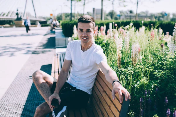 Pozitif gülümseyen genç bir erkek çiçek tarhının yanındaki parkta cep telefonu ve kulaklık kullanarak tek başına oturuyor.