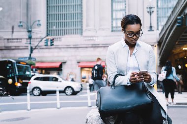 Şık giyinmiş, gözlüklü, yola yakın bir taşın üzerinde oturan ve şehir sokaklarının bulanık arka planında akıllı telefon kullanan etnik bir kadın.