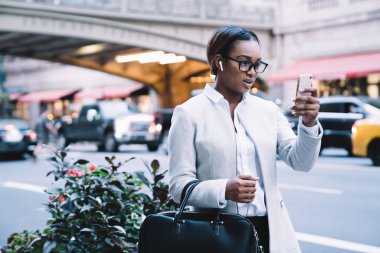 Beyaz, klasik takım elbiseli, kablosuz kulaklıklı, sokakta yürürken cep telefonundan izleyen dalgın, genç Afrikalı Amerikalı kadın.