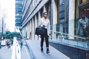 Ciddi Afro-Amerikalı siyah saçlı, gözlüklü, resmi giyimli, büyük siyah çantalı iş kadını gündüz vakti bulanık gökdelenlere karşı sokakta yürüyor.