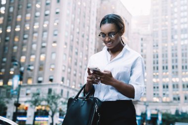 Gözlüklü, siyah çantalı ve resmi tişörtlü başarılı genç bir bayan iş gününün ardından iş bölgesinde telefon kullanarak arkadaşıyla mesajlaşıyor.