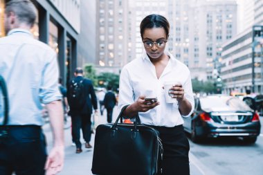 Modern odaklı Afro-Amerikan iş kadını resmi reklam bardakları içinde şehir merkezinde yürüyor bir fincan kahve içiyor ve akıllı telefondan mesajlaşıyor.