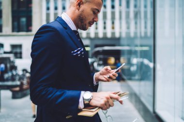 Düşük açılı yetişkin yönetici resmi giysileri içinde, ekranda yoğunlaşan belgelerle ve akıllı telefonla iletişim kuran şehir merkezindeki modern gökdelen binasının bulanık cephesine karşı caddede duran düşünceli yetişkin yöneticinin bakış açısı.