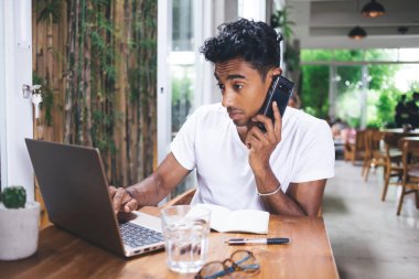 Beyaz tişörtlü Latin kökenli adam laptopunu kullanıyor ve kafeteryada bulanık arka planda otururken akıllı telefondan konuşuyor.