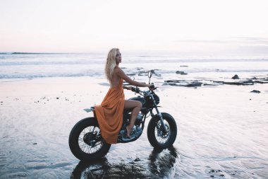 Açık turuncu elbiseli, kumlu sahil şeridi boyunca motosiklet süren ve kameraya bakan çekici sarışının yan görüntüsü. 