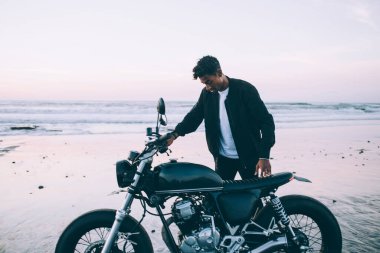 Sıradan giyinen genç İspanyol erkek motorcu motosikletin yanında durup boş kumlu sahil şeridinde at sürdükten sonra aşağı bakıyor.