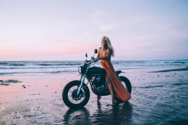 Siyah motosikletli, turuncu ışıklı seksi bir kadın nefes kesici su ve gökyüzünün arka planında gülümsüyor.