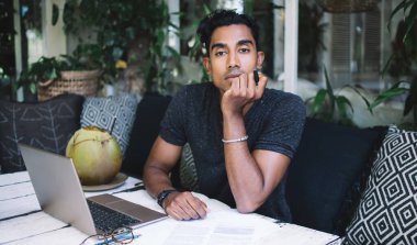 Zeki düşünceli İspanyol işadamı yeşil bitkilerle dolu bir kafede laptopla ve hindistan cevizi kokteyli içerken kalem ile kameraya bakıyor.